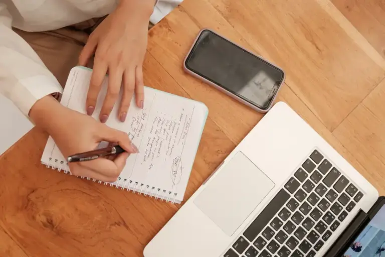 Person writing notes beside laptop and phone.